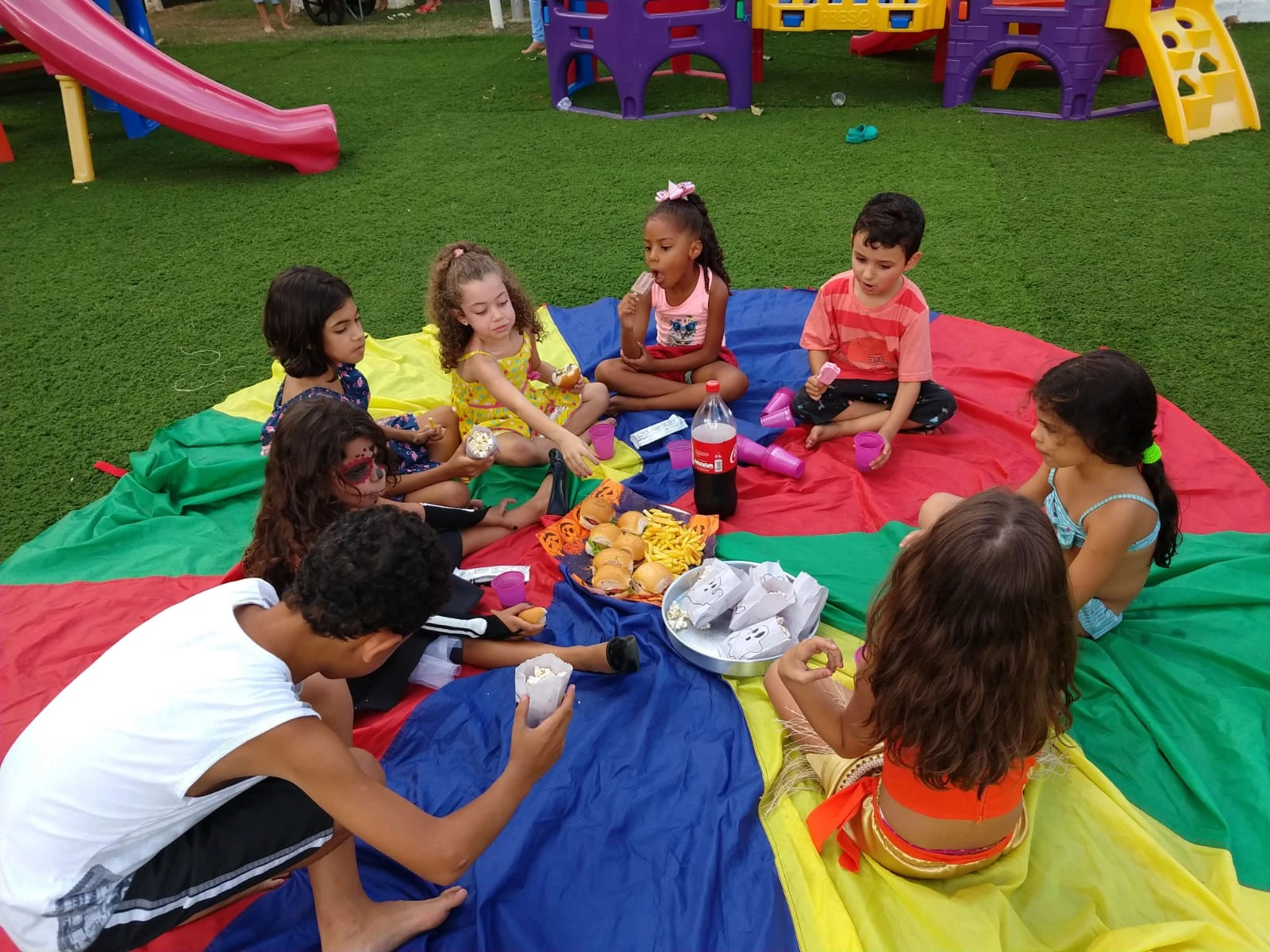 Crianças fazendo um piquenique sobre um paraquedas colorido no gramado do Espaço Kids